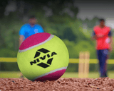 NIVIA Cricket Tennis Ball Light in neon green and pink resting on a cricket pitch surface, with the NIVIA logo visible in black. Two players and stumps appear blurred in the background, highlighting its use for cricket practice and recreational play.